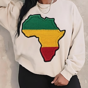 May include: Cream-colored sweatshirt featuring an embroidered map of Africa in green, yellow, and red. The sweatshirt has long sleeves and a crew neck. The design is centered on the front of the garment.