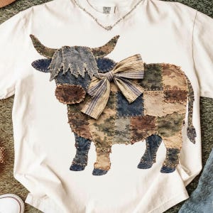 May include: Cream-colored t-shirt featuring a patchwork cow design. The cow is made of various fabrics in shades of blue, brown, and green, with a striped bow. The shirt has a distressed neckline and the text "CENTRAL COLORS".