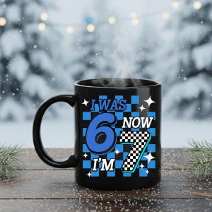 Pu&ograve; includere: Una tazza in ceramica nera con la scritta "I WAS 6 NOW I'M 7" in blu e bianco. Il numero 6 &egrave; in un grande carattere blu e il numero 7 ha un motivo a scacchiera. La tazza ha un manico nero.
