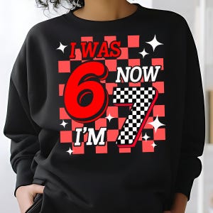 May include: Black sweatshirt with a red and white checkered design. The graphic reads "I WAS 6 NOW I'M 7" in a playful font. White star accents are scattered around the text, creating a celebratory theme.“six seven kid birthday”“i was six now i’m seven shirt”