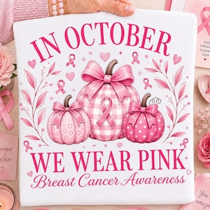 Op de afbeelding: Een wit t-shirt met een roze afbeelding van drie pompoenen met strikken, omringd door roze linten en bladeren. De tekst luidt: IN OCTOBER WE WEAR PINK Breast Cancer Awareness. De pompoenen hebben patronen van vichy, luipaardprint en stippen.
