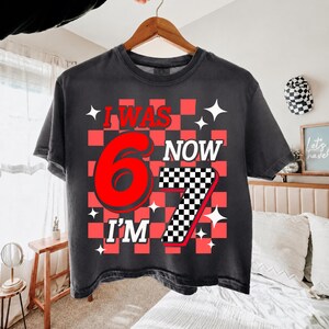 May include: A charcoal gray t-shirt featuring a red and black checkered background pattern. The graphic includes the red text I WAS, the large red number 6, the white text NOW, the white text I'M, and a large number 7 with a black and white checkerboard pattern.