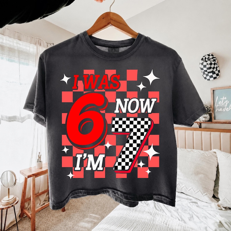 May include: A charcoal gray t-shirt featuring a red and black checkered background pattern. The graphic includes the red text I WAS, the large red number 6, the white text NOW, the white text I'M, and a large number 7 with a black and white checkerboard pattern.