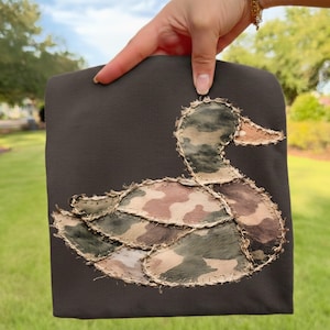 Op de afbeelding: Donkergrijs stoffen item met een eendenapplicatie. De eend is gemaakt van camouflage stof in groen, bruin en beige tinten, met een beige snavel. De applicatie is gestikt met een beige draad.