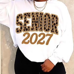 Op de afbeelding: Een wit sweatshirt met lange mouwen met het woord SENIOR in grote, luipaardprint chenille letters met een beige rand. Onder het woord staat het getal 2027 in grote, effen beige chenille cijfers met een bijpassende rand.