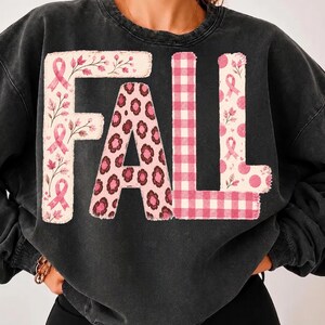 Op de afbeelding: Een antracietgrijze sweater met ronde hals en het woord FALL in grote blokletters. De F heeft roze linten en bladeren, de A een roze luipaardprint, de L een roze gingham-patroon en de tweede L roze stippen.