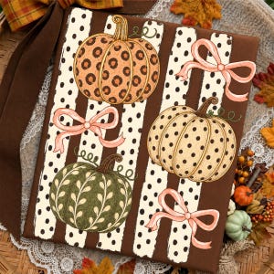 May include: A rectangular decorative sign featuring a brown background with vertical cream-colored stripes decorated with black polka dots. The design includes three illustrated pumpkins in orange leopard print, cream with black dots, and olive green with leaf patterns, accented by three light pink ribbons.
