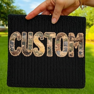 May include: Black knit beanie with the word "CUSTOM" embroidered in camouflage fabric. The letters are outlined with a tan-colored thread. The beanie is held up against a background of green grass and a blue sky.