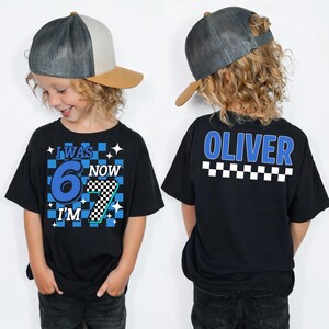 May include: Black t-shirt with the text "I WAS 6 NOW I'M 7" in blue and white, with a checkered pattern. The back of the shirt has the name "OLIVER" in blue, above a checkered pattern. The child is wearing a gray and tan baseball cap.