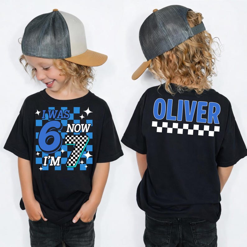 May include: Black t-shirt with the text "I WAS 6 NOW I'M 7" in blue and white, with a checkered pattern. The back of the shirt has the name "OLIVER" in blue, above a checkered pattern. The child is wearing a gray and tan baseball cap.