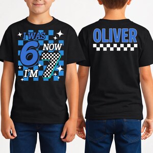 May include: Black t-shirt with the text "I WAS 6 NOW I'M 7" in blue and white, with a checkered pattern. The back of the shirt has the name "OLIVER" in blue.