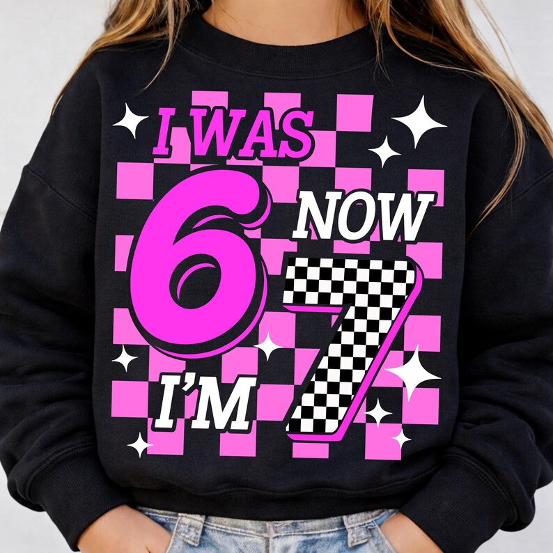 May include: Black sweatshirt with a pink and white checkered pattern. The text "I WAS 6 NOW I'M 7" is displayed in pink and white, with star accents. A fun birthday-themed design.