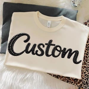 May include: Cream-colored t-shirt with the word "Custom" spelled out in black, beaded letters. The shirt is folded and resting on a gray pillow and a leopard print fabric.