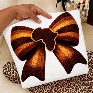 Peut inclure: Coussin carré blanc avec un grand motif de nœud papillon marron et doré orné d'une application en forme d'Afrique. Le nœud a un motif rayé et l'application est soulignée en or. Le coussin est sur une surface blanche avec du tissu à imprimé léopard.