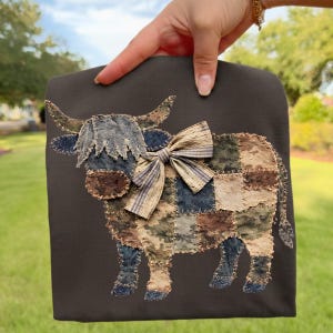 Könnte beinhalten: Eine dunkelbraune Tragetasche mit einem Patchwork-Kuh-Design. Die Kuh hat einen fransigen Denim-Look, eine gestreifte Schleife und Flicken aus Tarn- und Denim-ähnlichem Stoff. Die Hörner der Kuh sind hellbraun.