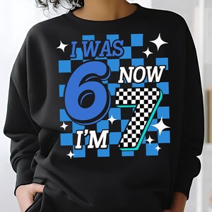 May include: Black sweatshirt with a blue and white checkered design. The graphic reads "I WAS 6 NOW I'M 7" in blue and white. White starbursts are scattered around the text. A fun birthday shirt.“i was six now i’m seven shirt” “six seven kid birthday”