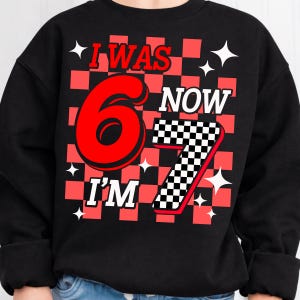 May include: A black long-sleeve sweatshirt featuring a graphic print with a red and black checkered background. The text reads I WAS 6 NOW I'M 7, with a large red number 6 and a checkered number 7. White four-pointed star shapes are scattered throughout.