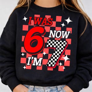 May include: A black crewneck sweatshirt featuring a graphic print with a red and black checkered background. The text reads I WAS 6 NOW I'M 7, with a large red number 6 and a black and white checkered number 7, accented by white star shapes.