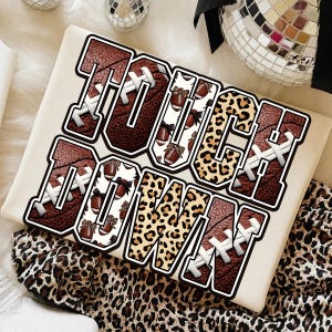 Op de afbeelding: Een wit t-shirt met de tekst TOUCHDOWN in grote blokletters. De letters zijn gevuld met patronen van bruine football-texturen, witte achtergronden met kleine footballs en strikjes, en een beige en zwart luipaardprintontwerp.