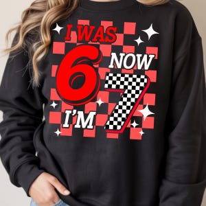 Puede incluir: Sudadera negra con un dise&ntilde;o a cuadros rojos y negros. El texto dice "I WAS 6 NOW I'M 7" en rojo y blanco. Acentos de estrellas blancas est&aacute;n dispersos alrededor del texto. Una divertida camiseta tem&aacute;tica de cumplea&ntilde;os.