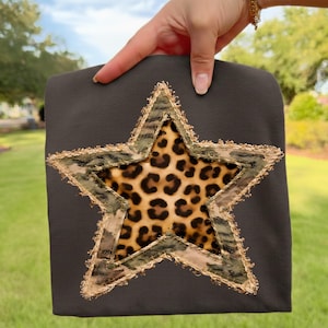 May include: A dark gray sweatshirt featuring a large star design. The star has a leopard print center, a camouflage border, and a frayed gold trim. The sweatshirt is held up against a green background.