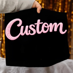 Puede incluir: Tela negra con la palabra "Custom" bordada en rosa. El bordado rosa tiene una apariencia texturizada. El fondo está borroso, con tonos dorados y blancos. La tela está siendo sostenida.