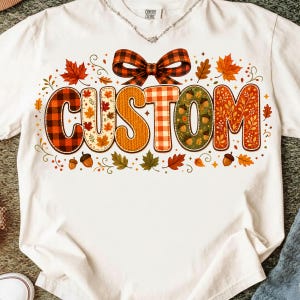 May include: A white short-sleeved t-shirt featuring the word CUSTOM in large, stylized letters. Each letter has a unique autumn-themed pattern, including plaid, leaves, and acorns. A large plaid bow sits above the text, surrounded by fall leaves.