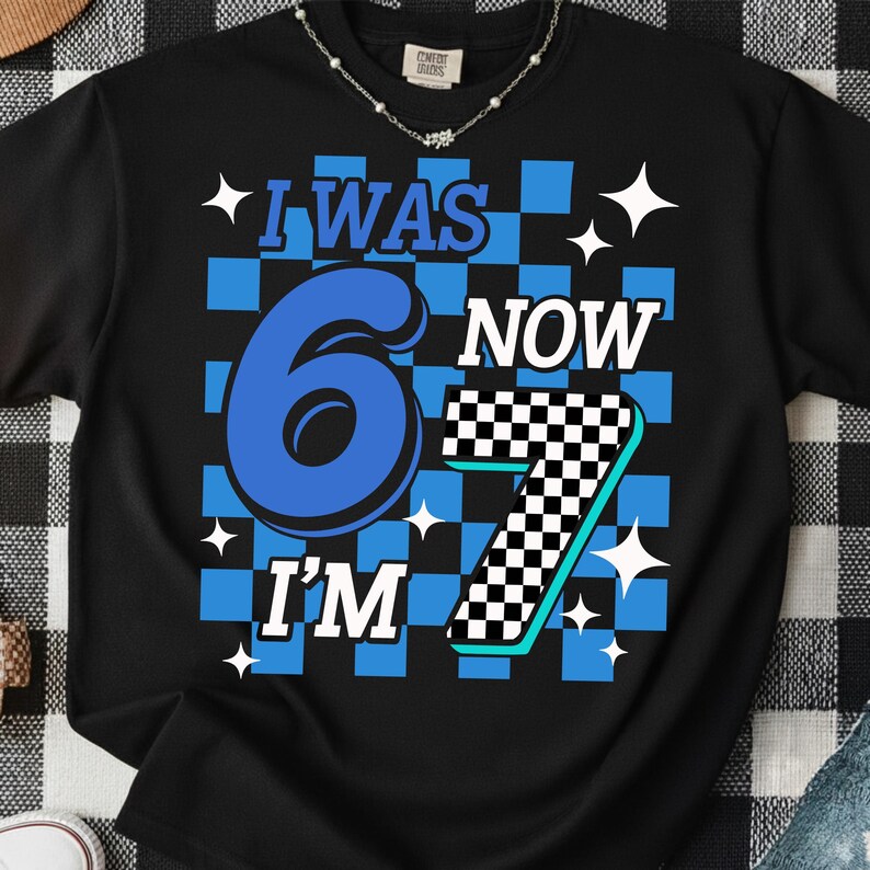 May include: Black t-shirt with a blue and white checkered design. The shirt features the text "I WAS 6 NOW I'M 7" in blue and white, with star accents. A fun birthday shirt.