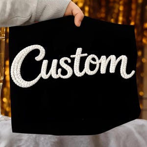 May include: A black fabric with the word "Custom" embroidered in white, held by a person. The text is in a cursive font. The background is blurred, with hints of gold and white.