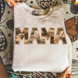 Puede incluir: Sudadera color crema con la palabra "MAMA" en letras grandes y texturizadas con estampado de camuflaje en la parte delantera. Las letras tienen un borde cosido de color dorado. La sudadera está doblada y sostenida por manos.