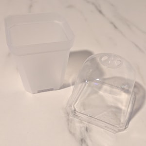 May include: A square, translucent, frosted plastic plant pot with a matching clear, dome-shaped plastic lid featuring a small circular ventilation opening at the top. The set consists of two separate pieces designed for small plant propagation.