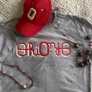 Può includere: Una maglietta grigia con la scritta "Ohio" in lettere rosse e bianche, un berretto da baseball rosso con una toppa "O" bianca e due collane con perline marroni e rosse. Gli articoli sono esposti su una superficie strutturata.