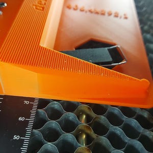 Könnte beinhalten: Nahaufnahme eines orangefarbenen Laserschneidwerkzeugs mit einem Lineal und einer schwarz-grauen Wabenoberfläche. Das Werkzeug hat eine geriffelte Oberseite und eine schwarze Komponente. Das Lineal zeigt Messungen von 50 bis 70.