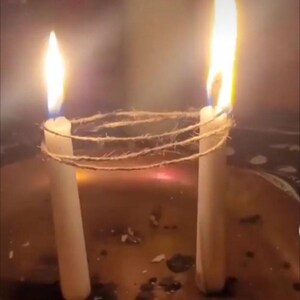 May include: Two white candles with bright yellow flames are connected by a twine. The candles are in a dark, possibly metallic container. Smoke rises around the candles, creating a mystical atmosphere.