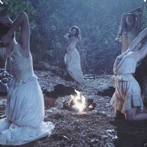 May include: A mystical scene depicts figures in vintage-style dresses gathered around a small bonfire in a forest. The dresses are light-colored, with lace and corset details. The setting is dimly lit, with a focus on the fire's glow.
