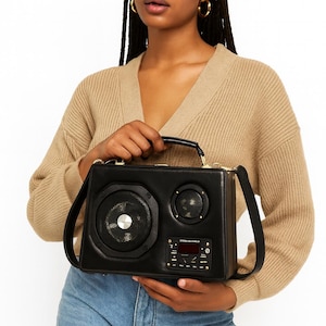 Puede incluir: Bolso de mano de cuero negro dise&ntilde;ado para parecerse a un boombox vintage. El bolso presenta detalles de altavoces, un asa y una correa para el hombro. El panel frontal incluye una pantalla digital y botones de control. El bolso lo sostiene una persona con gafas de sol y un c&aacute;rdigan beige.