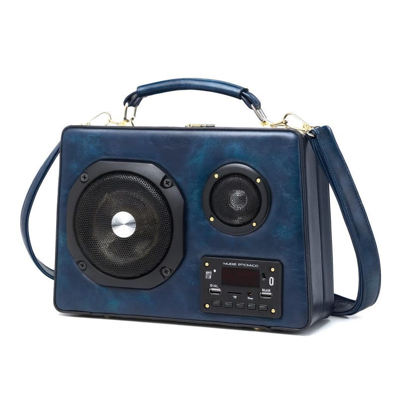 Puede incluir: Un bolso azul marino dise&ntilde;ado para parecerse a una radio vintage. El bolso cuenta con dos altavoces, una pantalla digital y botones de control. Tiene un asa superior y una correa para el hombro desmontable. El bolso est&aacute; hecho de un material texturizado.