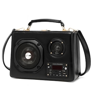 Puede incluir: Bolso de mano negro de cuero sint&eacute;tico dise&ntilde;ado para parecerse a un boombox vintage. Cuenta con un altavoz grande, un altavoz m&aacute;s peque&ntilde;o y una pantalla digital. El bolso tiene un asa superior y una correa para el hombro. El texto "MUSIC SPEAKERS" es visible.