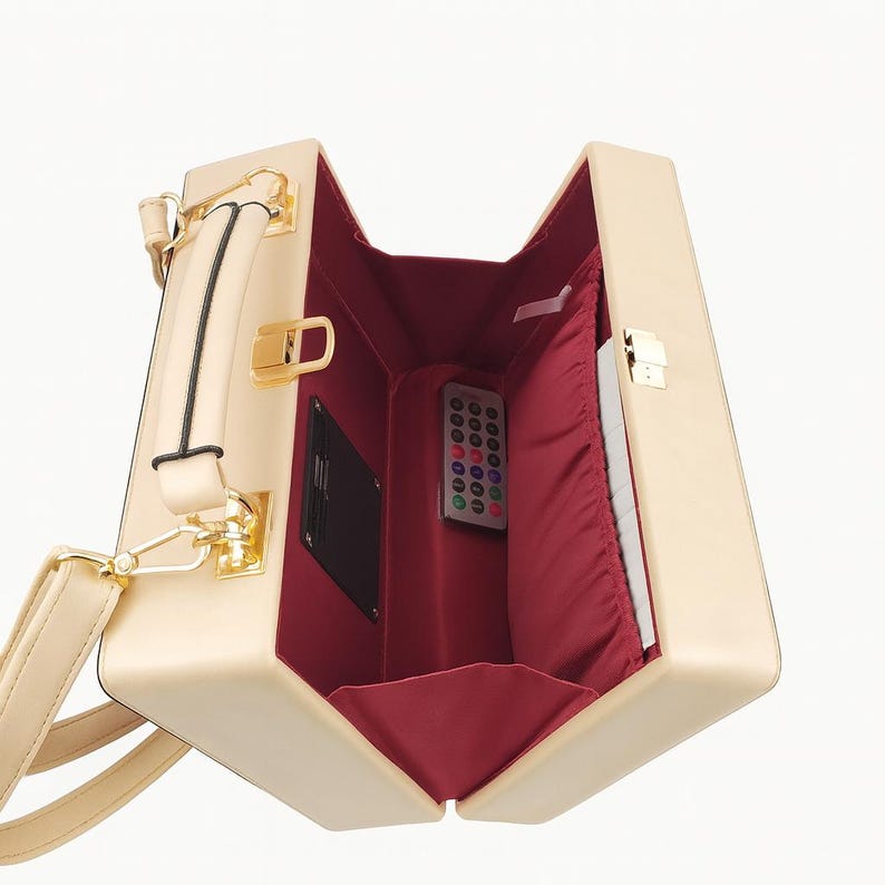Puede incluir: Bolso de mano rectangular beige con cierre y asa dorados. El interior est&aacute; forrado con tela roja y contiene un mando a distancia y una peque&ntilde;a tarjeta negra. El bolso tiene una correa para el hombro desmontable.