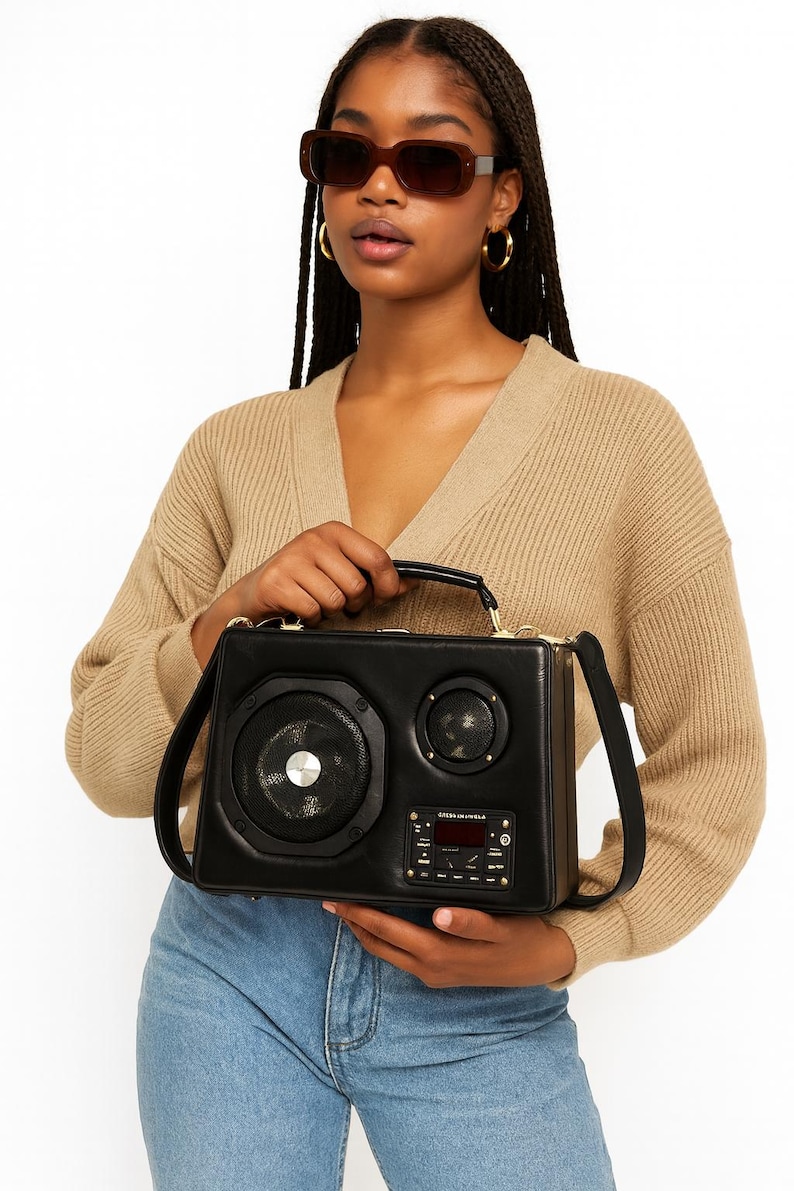 Puede incluir: Bolso de mano de cuero negro dise&ntilde;ado para parecerse a un boombox vintage. El bolso presenta detalles de altavoces, un asa y una correa para el hombro. El panel frontal incluye una pantalla digital y botones de control. El bolso lo sostiene una persona con gafas de sol y un c&aacute;rdigan beige.
