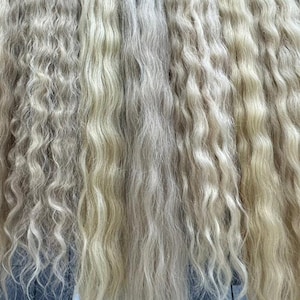 以下が含まれることがあります： ウェーブのかかったブロンドのヘアエクステンションの展示。暗いベースの各束は、金属製のラックに吊り下げられています。さまざまなブロンドの色合いと髪のウェーブのかかった質感がはっきりと見えます。