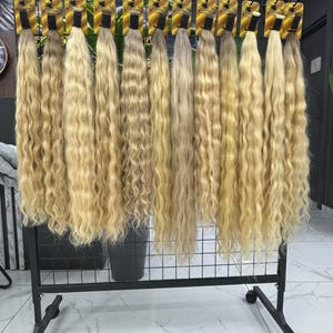 以下が含まれることがあります： 黒い金属ラックに吊るされた、ブロンドのウェーブのかかったヘアエクステンションのディスプレイ。髪は明るいブロンドからミディアムブロンドまで様々な色合いで、自然なウェーブがかかっています。各束は小さなカードに付いています。