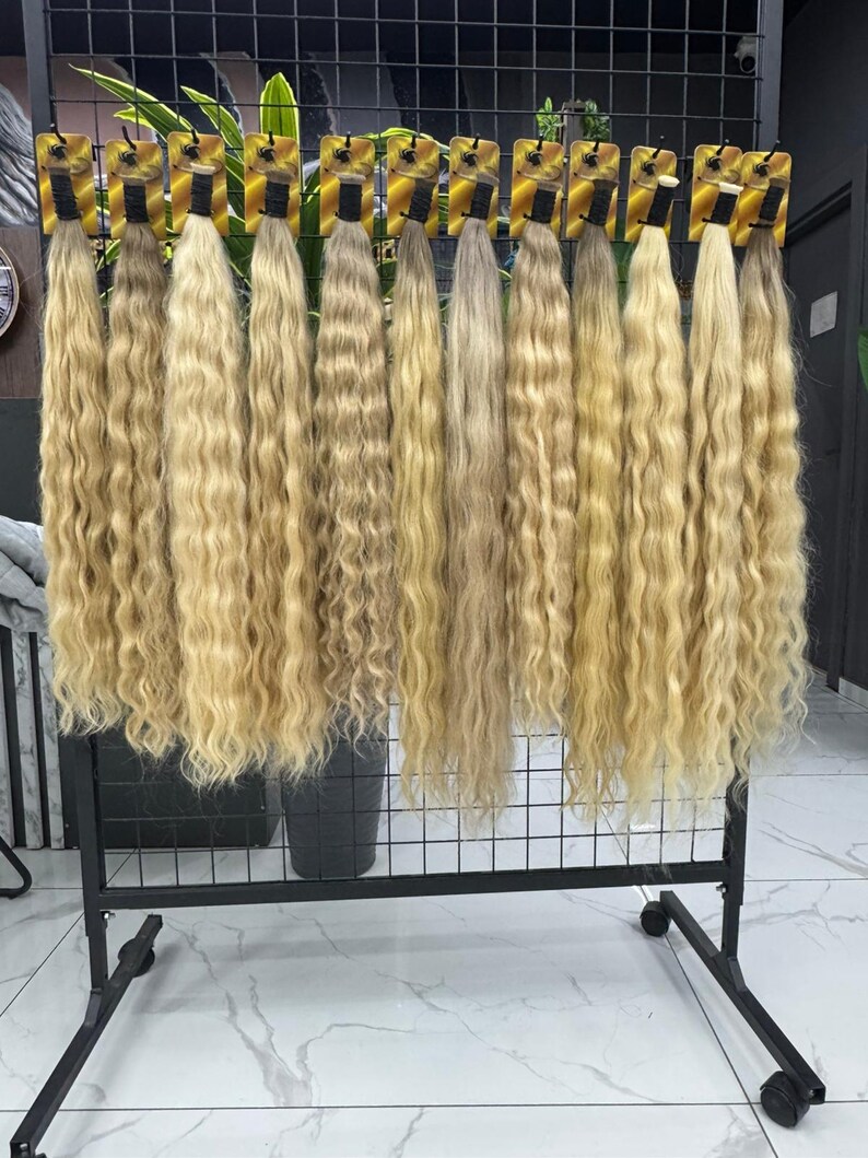 以下が含まれることがあります： 黒い金属ラックに吊るされた、ブロンドのウェーブのかかったヘアエクステンションのディスプレイ。髪は明るいブロンドからミディアムブロンドまで様々な色合いで、自然なウェーブがかかっています。各束は小さなカードに付いています。