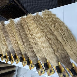 以下が含まれることがあります： 白い表面に並べられた、波状の明るいブロンドのヘアエクステンションの束がいくつかあります。髪は自然な波模様とグラデーション効果があり、暗い根元から明るい毛先へと変化します。各束は黒いバンドで固定されています。