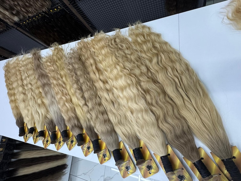 以下が含まれることがあります： 白い表面に並べられた、波状の明るいブロンドのヘアエクステンションの束がいくつかあります。髪は自然な波模様とグラデーション効果があり、暗い根元から明るい毛先へと変化します。各束は黒いバンドで固定されています。