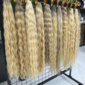 以下が含まれることがあります： 黒い金属製のラックに吊るされた、ブロンドのウェーブのかかったヘアエクステンションのディスプレイ。髪は明るい色から中程度の色まで、さまざまなブロンドの色合いがあります。各バンドルは、上部に黒いバンドと黄色のタグで固定されています。