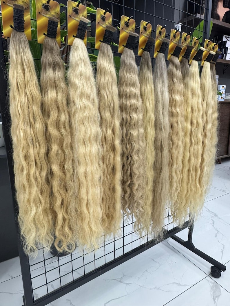 以下が含まれることがあります： 黒い金属製のラックに吊るされた、ブロンドのウェーブのかかったヘアエクステンションのディスプレイ。髪は明るい色から中程度の色まで、さまざまなブロンドの色合いがあります。各バンドルは、上部に黒いバンドと黄色のタグで固定されています。