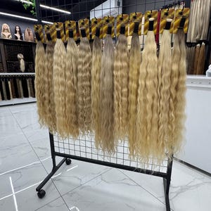 以下が含まれることがあります： さまざまな色合いのウェーブのかかったブロンドのヘアエクステンションが、黒い金属製のラックに吊り下げられています。エクステンションは小さなカードに付けられており、さまざまな色と質感を展示するように並べられています。