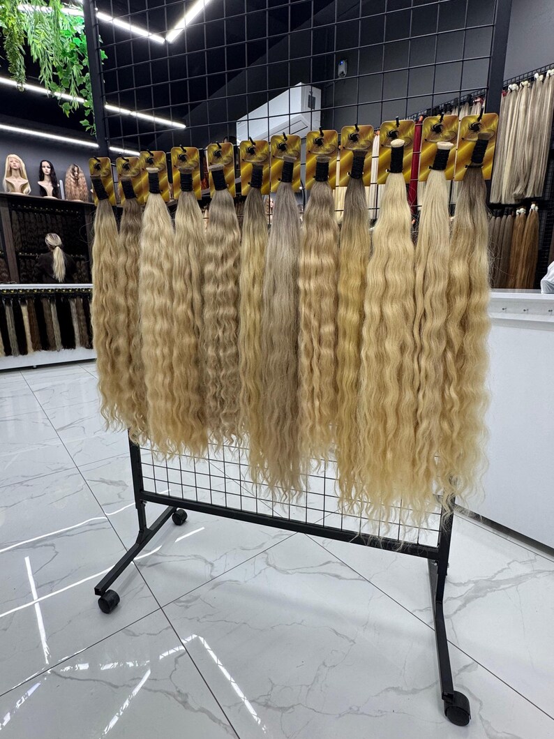 以下が含まれることがあります： さまざまな色合いのウェーブのかかったブロンドのヘアエクステンションが、黒い金属製のラックに吊り下げられています。エクステンションは小さなカードに付けられており、さまざまな色と質感を展示するように並べられています。