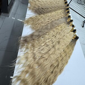以下が含まれることがあります： 白い表面に並べられた、波状の明るいブロンドのヘアエクステンションの束がいくつかあります。髪は自然で質感があり、さまざまなブロンドの色合いがあります。各束は上部で黒いバンドで結ばれています。